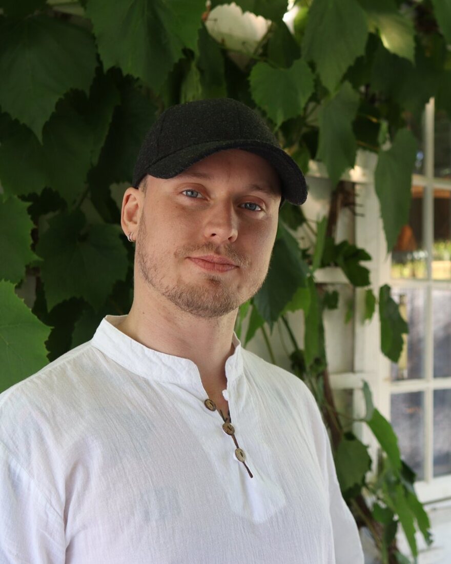 Fritz Himberg i hvit skjorte og svart caps foran en vegg med vinranker